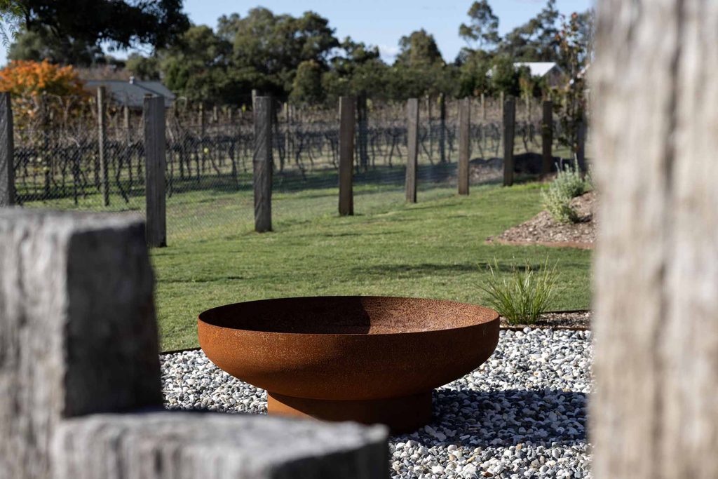 Corten steel firepit bowl framed by timber posts, with vineyard rows visible in the background