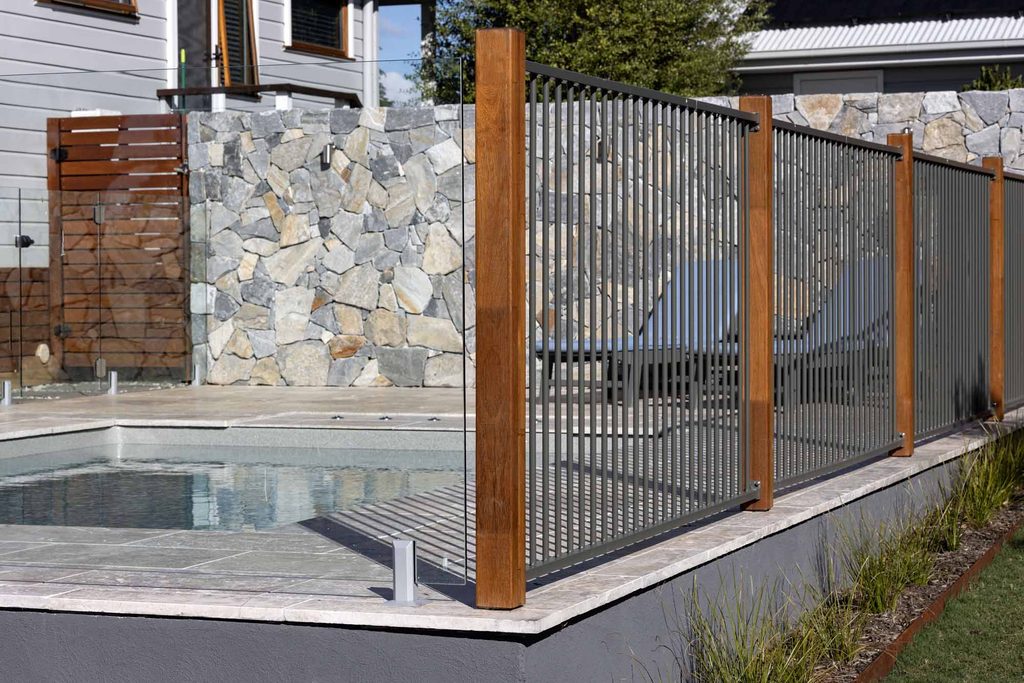 Glass pool fencing panel with merbau timber posts and aluminium slat fencing against a stone wall